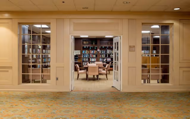 View through open double doors into a library room with bookshelves filled with books and a round table surrounded by six chairs. The room is framed by beige walls with large windows on either side of the doors, and a patterned carpet in the foreground.