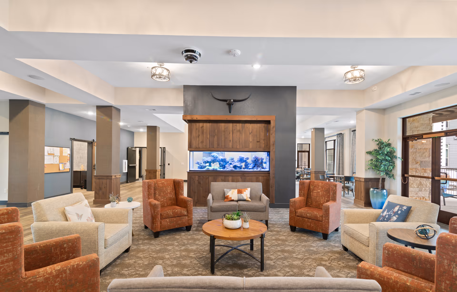 A spacious and well-lit living room area in a senior living facility featuring a circular seating arrangement with beige and rust-colored armchairs around a wooden coffee table. A large built-in aquarium is set into a dark accent wall with a decorative longhorn skull mounted above it. The room has light-colored walls, wood flooring, and large windows letting in natural light. There are ceiling lights and a potted plant near the entrance door.