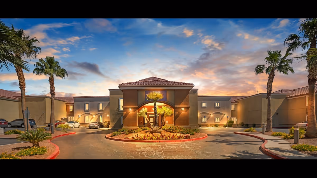 Front entrance of a two-story senior living facility with a circular driveway, palm trees, and a colorful sunset sky.