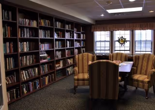 A cozy library room with tall wooden bookshelves filled with books along one wall. There is a table surrounded by four upholstered armchairs with striped fabric. Three windows with blinds and a valance allow natural light into the room.