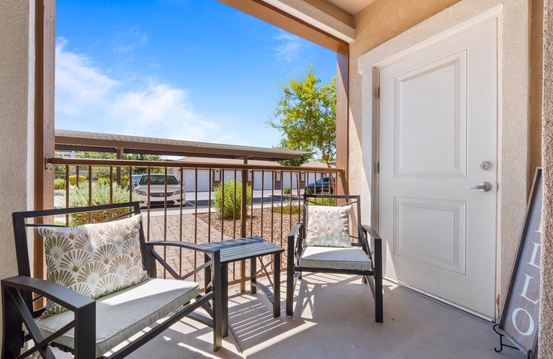 A small outdoor patio area with two black metal chairs featuring patterned cushions and a small matching table between them. The patio is enclosed by a metal railing and overlooks a driveway with parked cars and a tree under a clear blue sky.