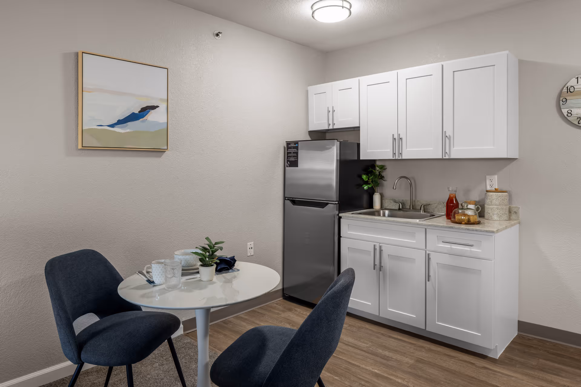 A small kitchen area with white cabinets, a stainless steel refrigerator, a sink, and a countertop with a glass pitcher and cups. In front of the kitchen is a small round white table with two dark blue upholstered chairs. A small plant and two cups are on the table. The walls are light-colored with a framed abstract painting and a round wall clock.