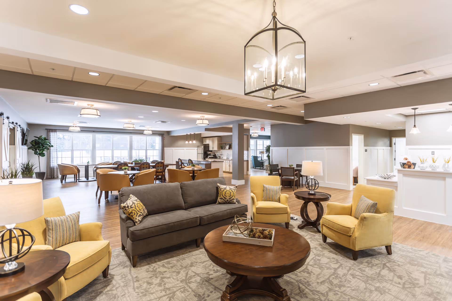 A spacious and well-lit senior living common area featuring a gray sofa, three yellow armchairs, round wooden coffee tables, and a large area rug. In the background, there are multiple dining tables and chairs near large windows letting in natural light. The room has modern light fixtures and a neutral color palette with wood flooring.