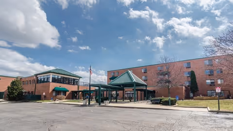 Exterior view of a senior living facility named Heartland Of Woodridge featuring a large brick building with multiple windows, green roofs over entrances, a covered drop-off area, a flagpole with an American flag, and a parking lot in the foreground under a partly cloudy sky.