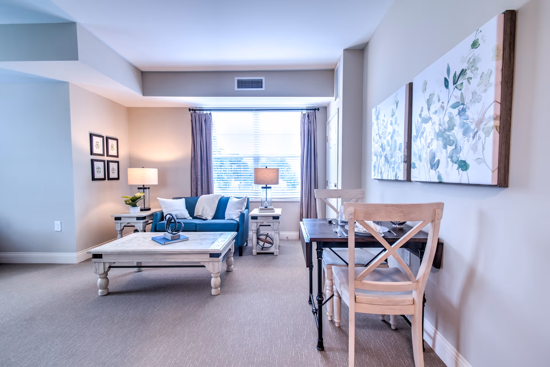 Bright furnished living room with a blue sofa, coffee table, side tables, lamps and a small dining/desk area by a window.