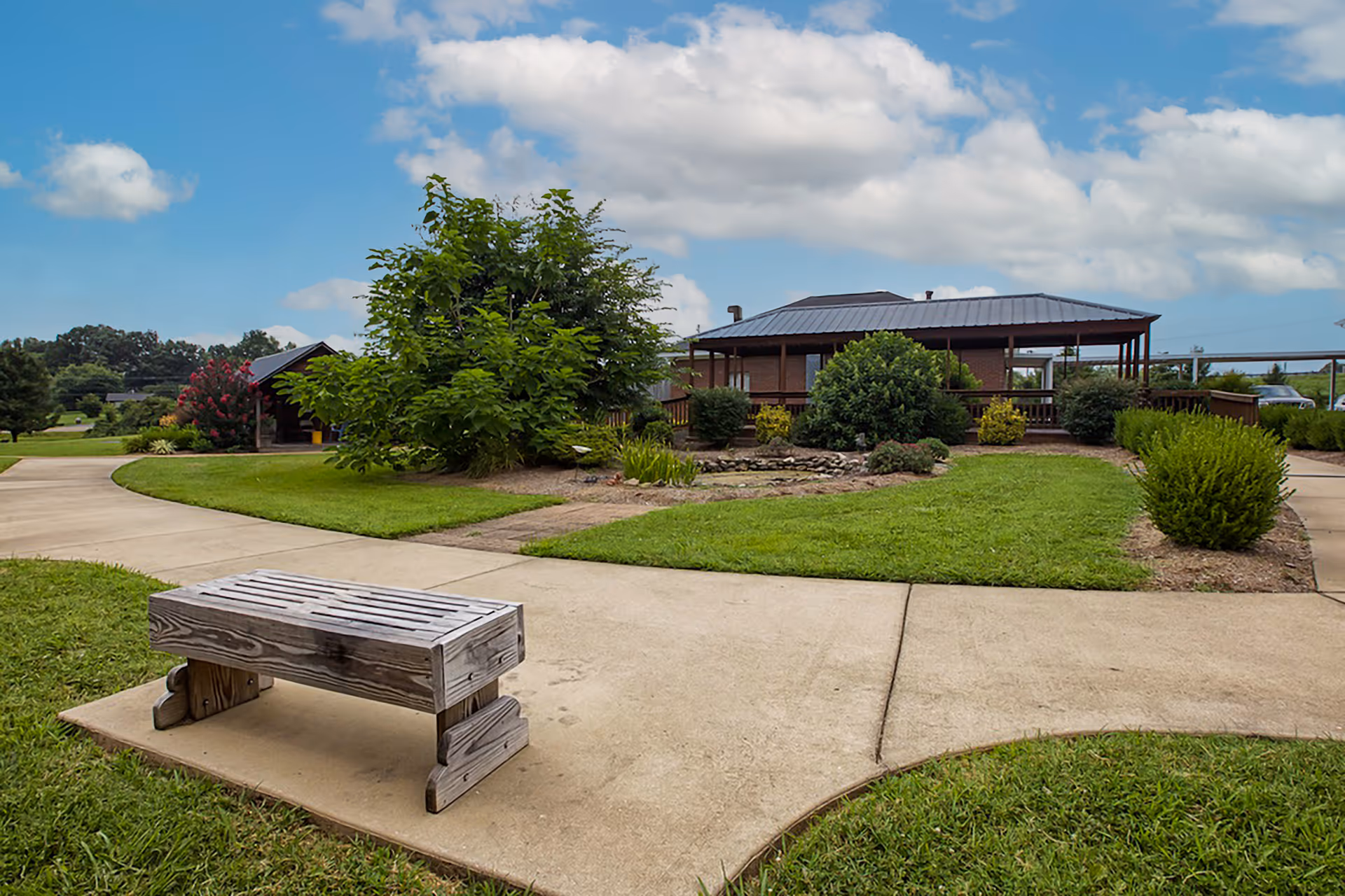 Outdoor area with a wooden bench on a concrete pathway surrounded by green grass, bushes, and trees. In the background, there is a single-story building with a metal roof and a covered porch. The sky is partly cloudy.