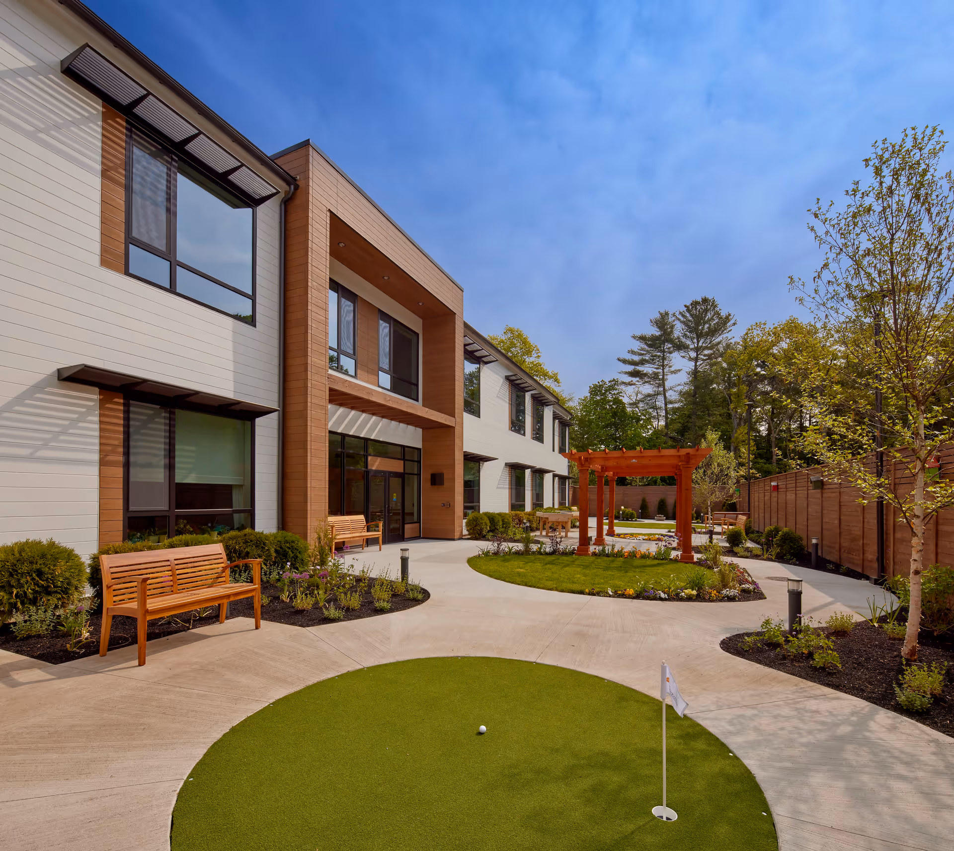 Outdoor courtyard area of The Cordwainer facility featuring a small putting green with a golf ball and flag, wooden benches, landscaped flower beds, a wooden pergola, and a two-story building with large windows under a blue sky.