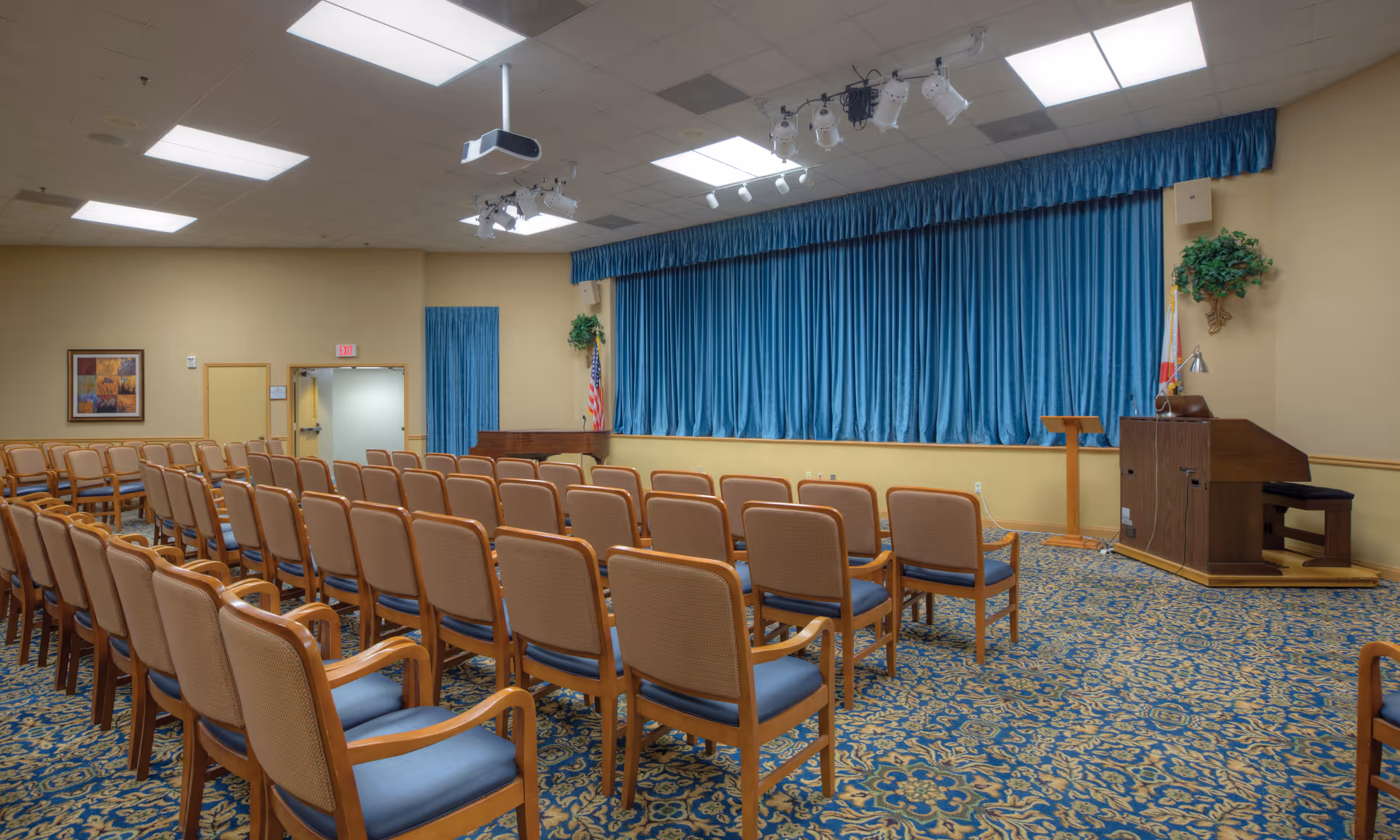 An empty meeting or event room with rows of wooden chairs with blue cushions facing a stage with blue curtains. The room has a patterned blue and beige carpet, beige walls, ceiling lights, a projector, and a podium with a microphone. There are two flags and some greenery on the stage area.