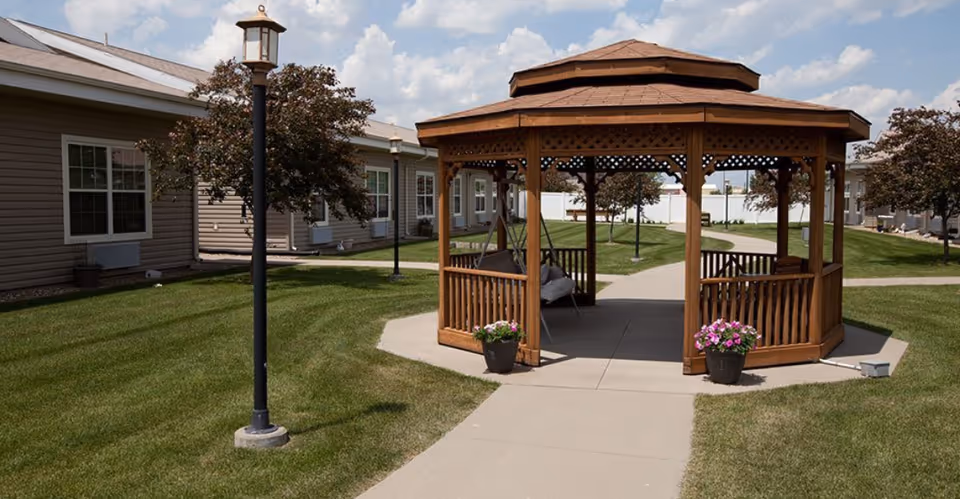 A wooden gazebo sits on a paved path in a grassy courtyard between single-story assisted living buildings.