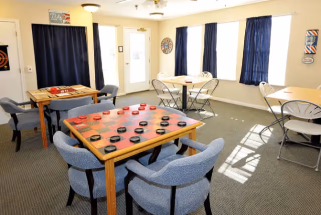 A bright room with several tables and chairs arranged for games and social activities. One table has a large checkers board with checkers pieces set up, and another table has a backgammon board. The room has windows with blue curtains, a dartboard on the wall, and a sign that reads 'Barber Shop'.