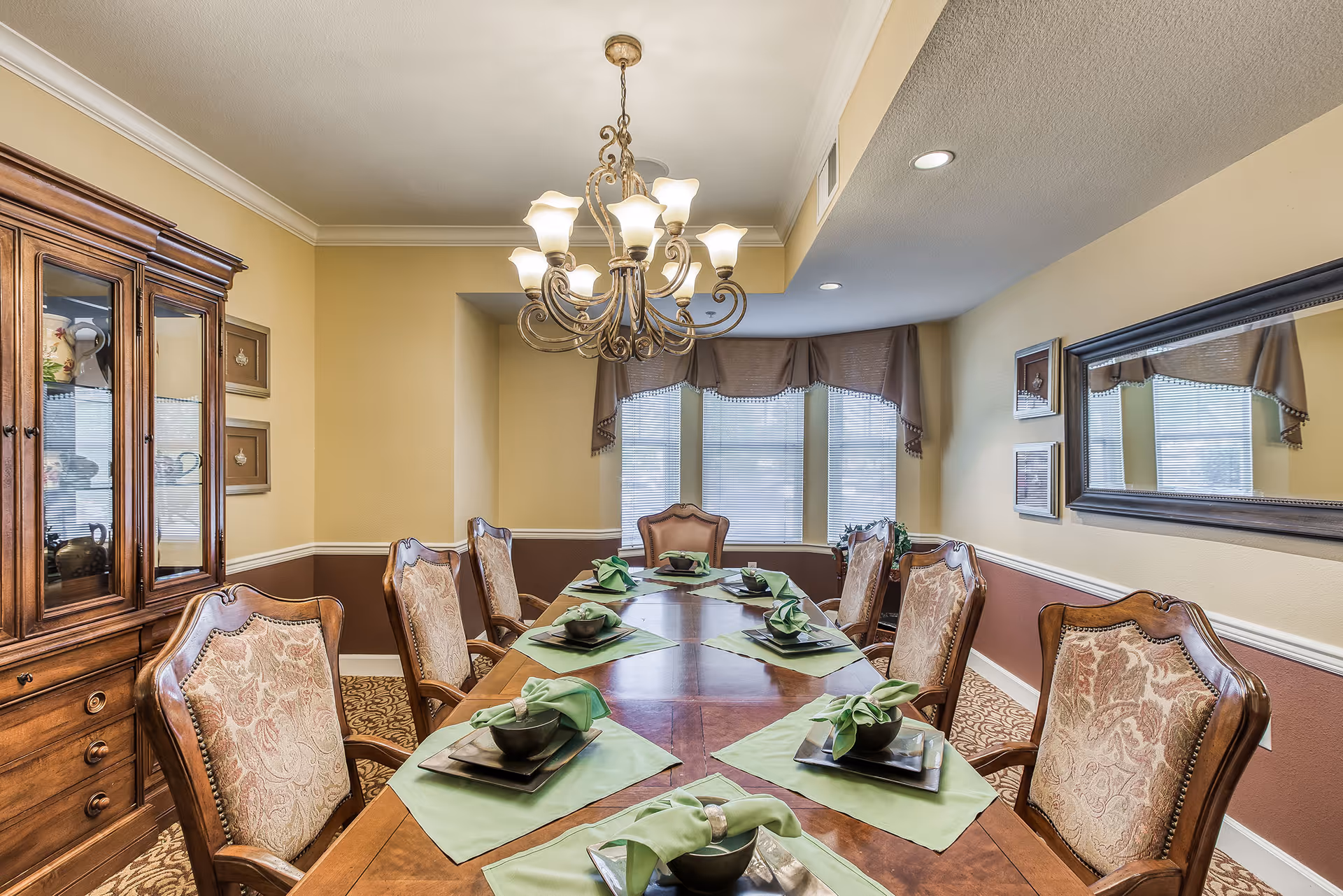 A formal dining room with a long wooden table set with green napkins and black bowls on square plates. The room features upholstered wooden chairs, a large chandelier hanging from the ceiling, a wooden cabinet with glass doors, framed artwork on the walls, and a large mirror reflecting the window with brown curtains.