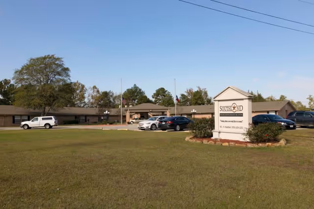 Exterior view of Southland Rehabilitation And Healthcare Center showing a single-story building with a parking lot in front, several cars parked, a grassy lawn, and a sign with the facility's name. Trees and a clear blue sky are visible in the background.