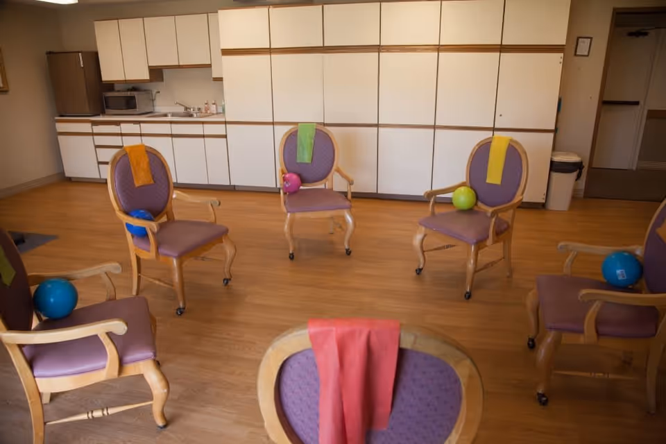 A room with six wooden chairs arranged in a circle, each chair has a colored towel draped over the back and a small exercise ball placed on the seat. The room has wooden flooring and a wall with white cabinets and a countertop with a microwave and sink.