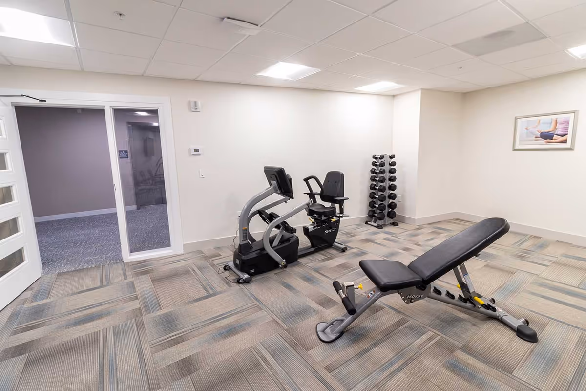 A small fitness room with a recumbent exercise bike, an adjustable weight bench, and a rack of dumbbells. The room has light-colored walls, a patterned carpet floor, a framed picture on the wall, and a glass door leading to another area.