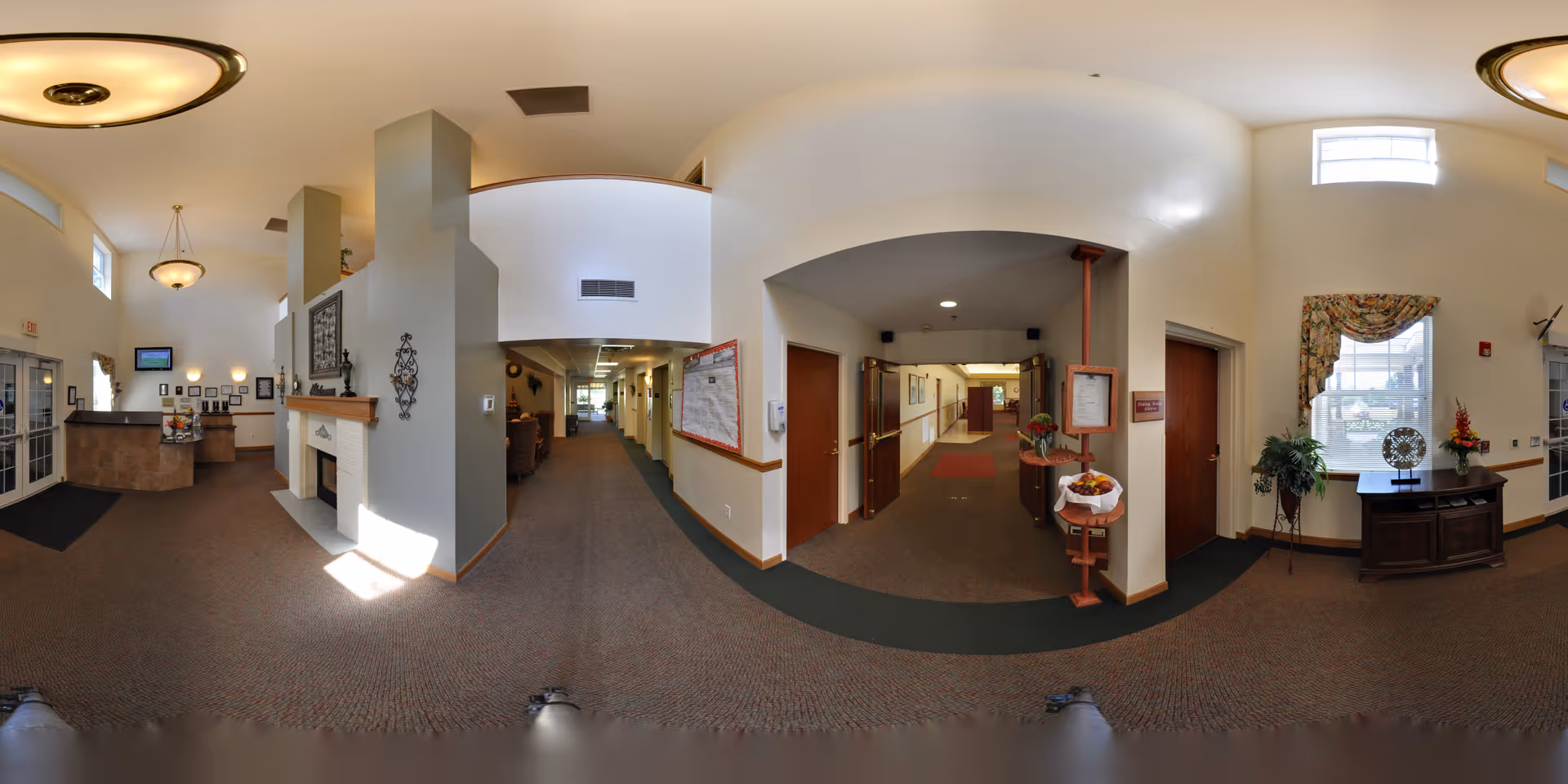 Spacious senior living facility lobby with a reception desk, fireplace, seating areas and a central hallway leading to rooms.