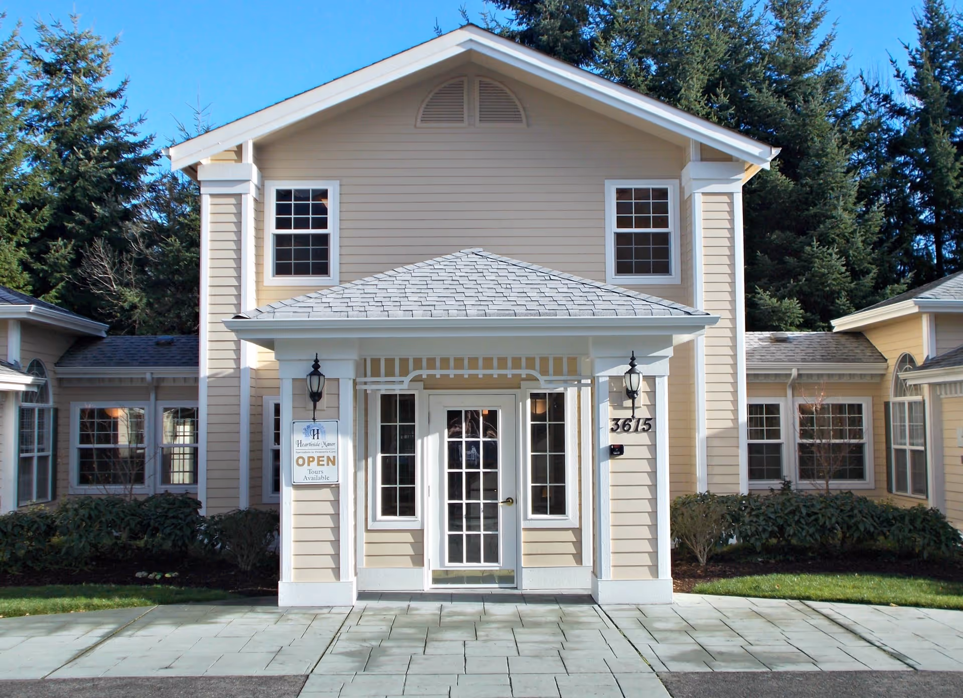 Front exterior view of Hearthside Manor Memory Care building with beige siding, white trim, and a covered entrance with glass double doors. There are two black lantern-style lights on either side of the entrance and a sign indicating the facility is open with tours available. The building is surrounded by greenery and trees under a clear blue sky.
