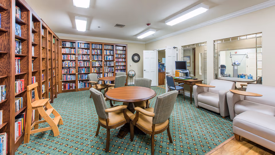 A well-lit senior living library/common room with wall-to-wall bookshelves, round tables and chairs, and lounge seating.
