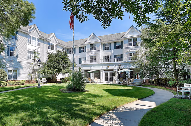 White multi-story senior living building with manicured lawn, walkway, and outdoor seating.