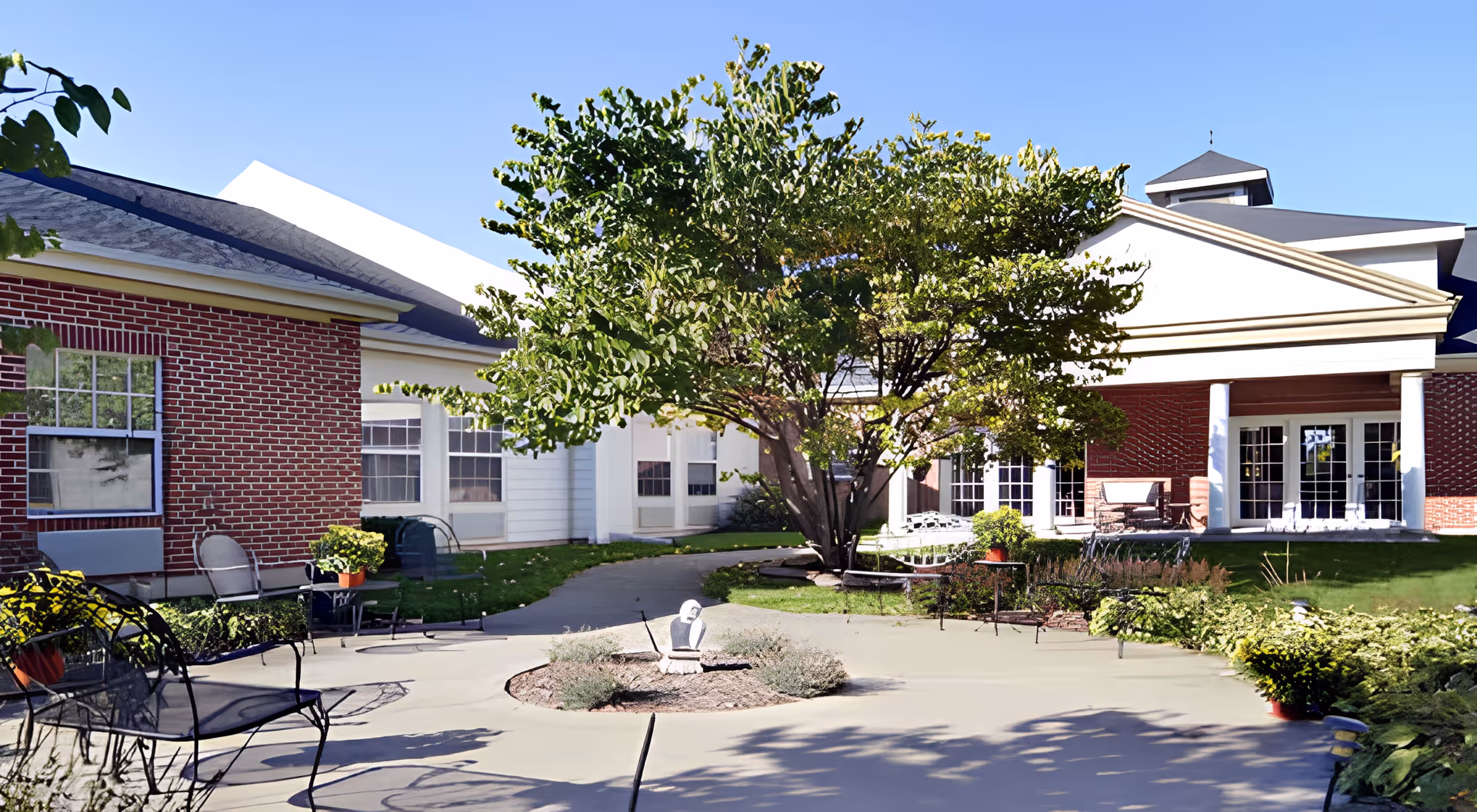 Outdoor courtyard area of a senior living facility with a large tree in the center, surrounded by a paved walkway, garden beds, and outdoor seating including metal chairs and tables. The building has red brick and white siding with large windows and a covered patio area.