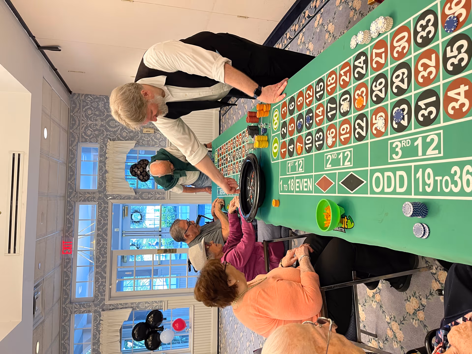 A group of elderly people sitting and standing around a green roulette table inside a room with patterned carpet and wallpaper. A man with a beard and wearing a white shirt and black vest is operating the roulette wheel while the others watch and participate. There are balloons in the background near a glass door with an exit sign above it.
