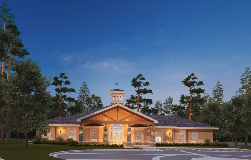 Exterior front view of a single-story building with a peaked roof and a central entrance featuring large glass doors and stone pillars, surrounded by trees and a clear evening sky.