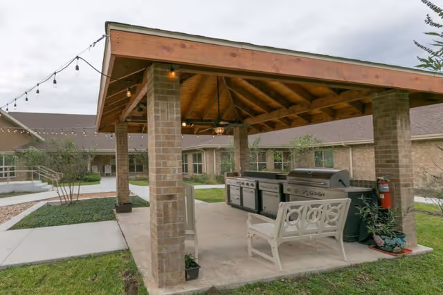 Outdoor covered patio area with a wooden roof supported by brick pillars, featuring a large grill and cooking station, a white bench, string lights hanging from the roof, and surrounding greenery with a building in the background.