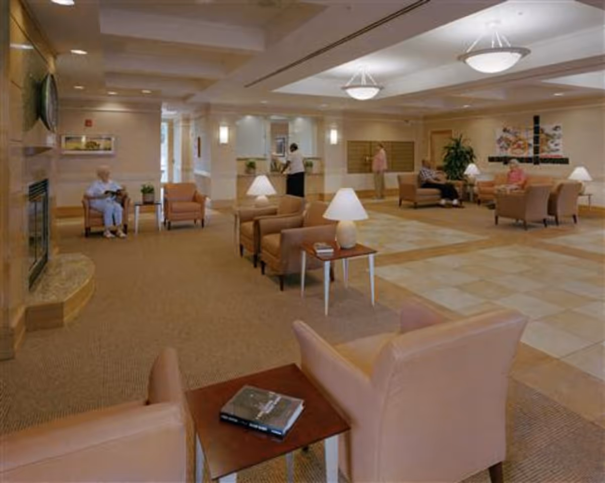 A spacious senior living facility common area with beige walls and carpeted floor. Several comfortable armchairs and small tables with lamps are arranged throughout the room. There are a few elderly people sitting and standing, engaging in quiet activities. The room is well-lit with ceiling lights and has a welcoming atmosphere.