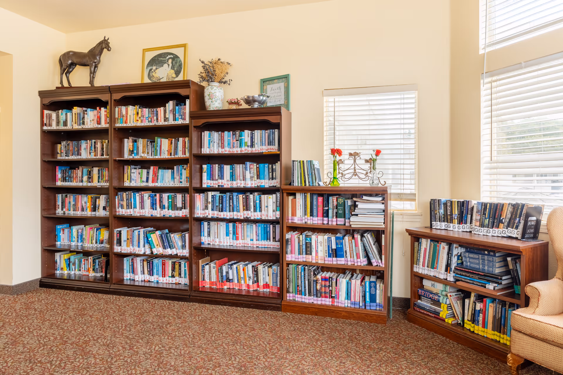 A cozy interior reading area with several wooden bookshelves filled with books beside two windows and a cushioned chair.