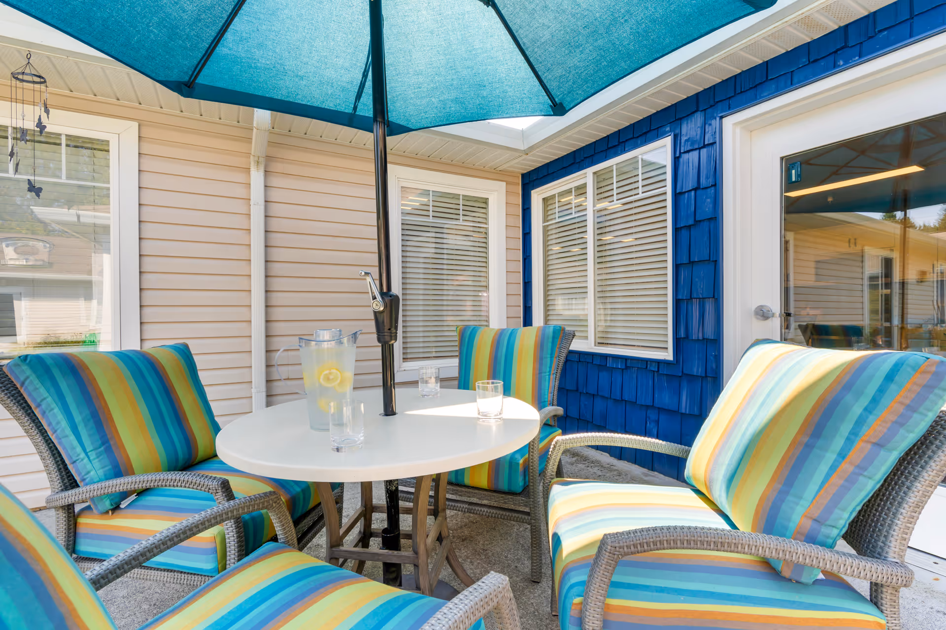 Outdoor patio area with a round white table and four wicker chairs with colorful striped cushions. A large blue umbrella provides shade over the table, which has a pitcher of lemon water and three glasses. The patio is adjacent to a building with beige and blue siding and several windows.