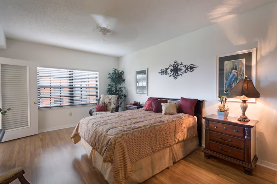 A well-lit bedroom with a large bed covered in a patterned beige and brown bedspread with multiple pillows. There is a wooden nightstand with a lamp and a small plant on the right side of the bed. A framed picture of a bird hangs on the wall above the nightstand. On the left side of the room, there is a window with blinds and a cushioned armchair with a pillow next to a small table. The floor is wooden, and the walls are painted white.