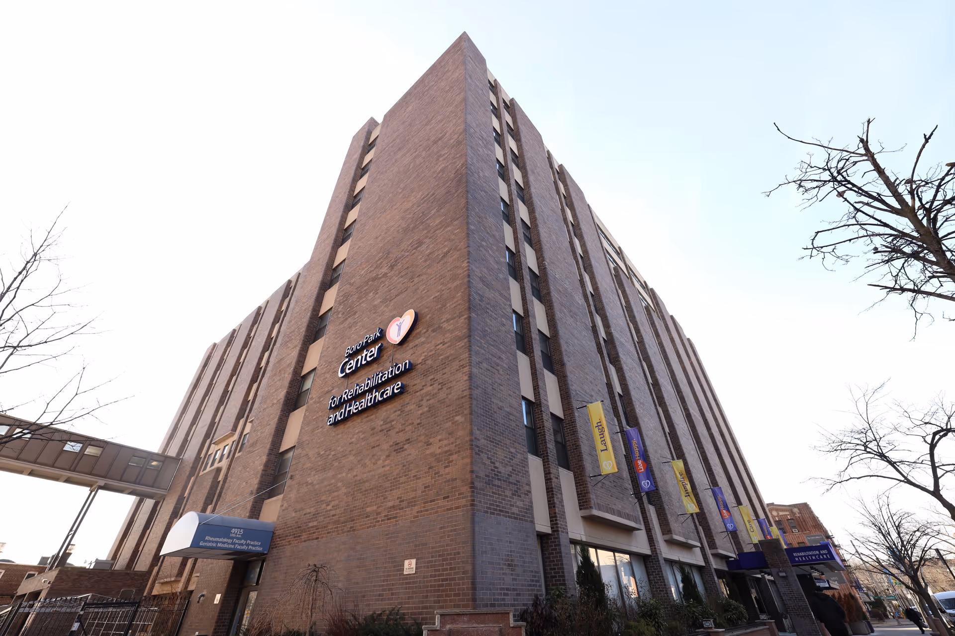 Exterior view of a multi-story brick building with a sign that reads 'Boro Park Center for Rehabilitation and Healthcare'. The building has several windows and banners hanging on the side. Leafless trees are visible in the foreground under a clear sky.
