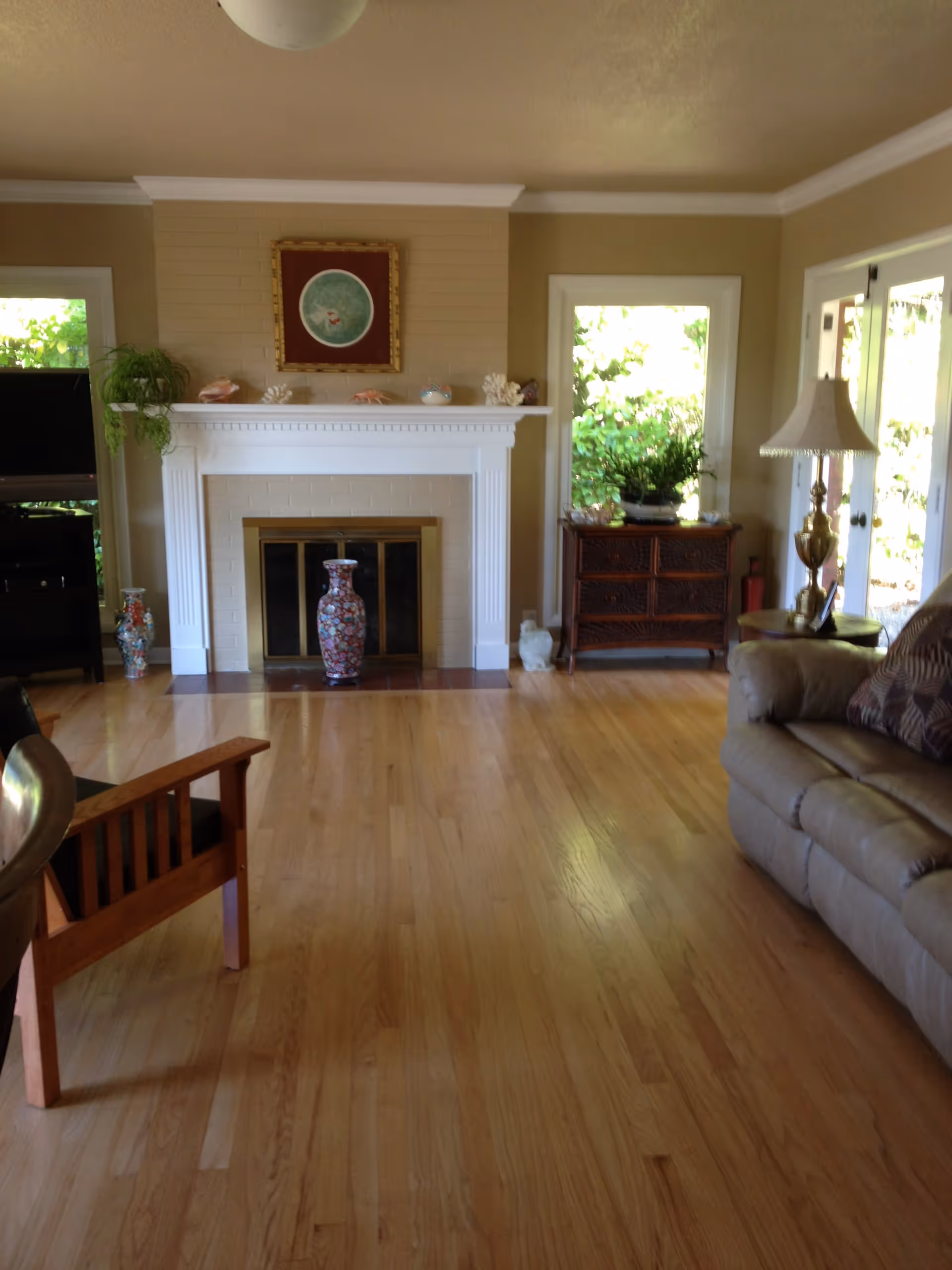 A cozy living room featuring a fireplace with decorative items on the mantel, a wooden chair, and a comfortable sofa. Large windows allow natural light to fill the space, showcasing a wooden floor and a warm color palette.