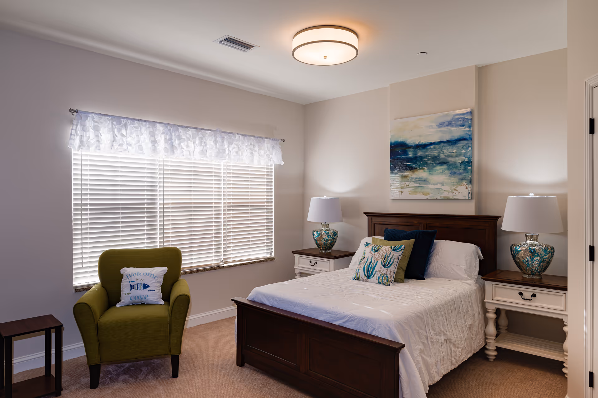 A cozy bedroom with a wooden bed frame, white bedding, and decorative pillows. There are two matching bedside tables with ornate lamps on each side of the bed. A green armchair with a decorative pillow sits near a window with white blinds and a sheer valance. A large abstract painting with blue and green tones hangs above the bed.