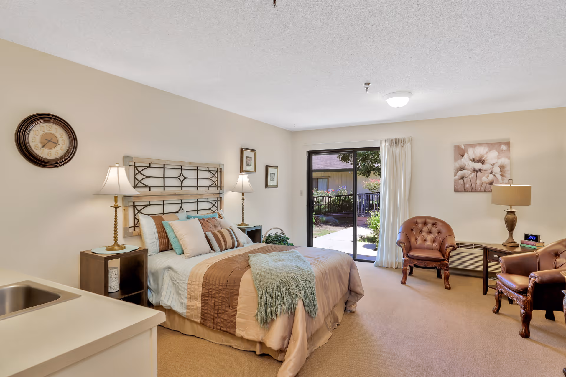 A well-lit bedroom in an assisted living facility featuring a queen-sized bed with multiple pillows and a throw blanket. The room includes two bedside tables with lamps, a wall clock, two armchairs, a side table with a lamp and clock, and a sliding glass door leading to an outdoor garden area.