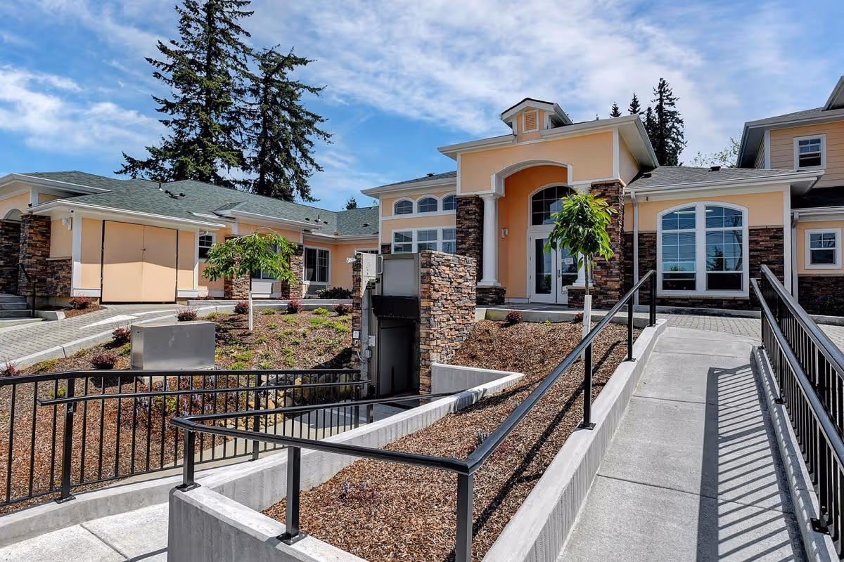 Exterior view of Tabor Crest Memory Care facility showing a peach-colored building with stone accents, large windows, and a main entrance with double doors. There is a concrete wheelchair ramp with black railings leading up to the entrance, landscaped areas with small trees and shrubs, and a clear blue sky with some clouds in the background.