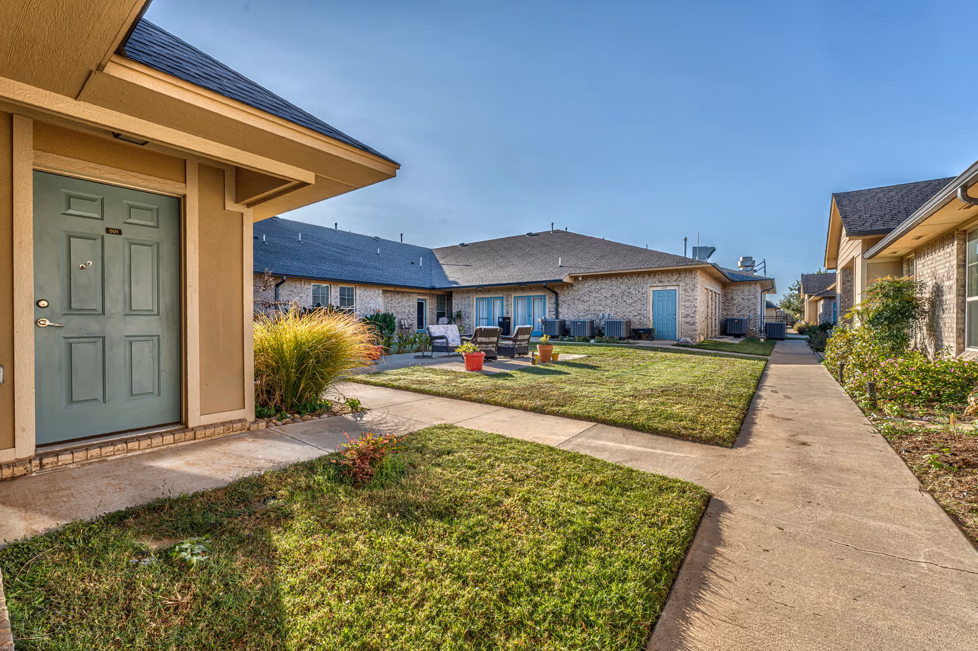 Outdoor view of a senior living facility courtyard with beige brick buildings, green doors, a well-maintained lawn, paved walkways, and outdoor seating with chairs and potted plants under a clear blue sky.