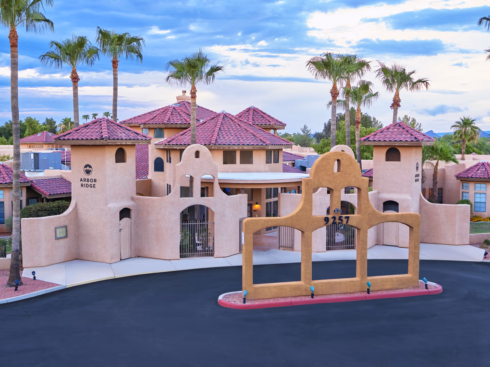 Exterior view of Arbor Ridge senior living facility with southwestern-style architecture, featuring beige stucco walls, red tile roofs, palm trees, and a decorative entrance structure with the number 9257.