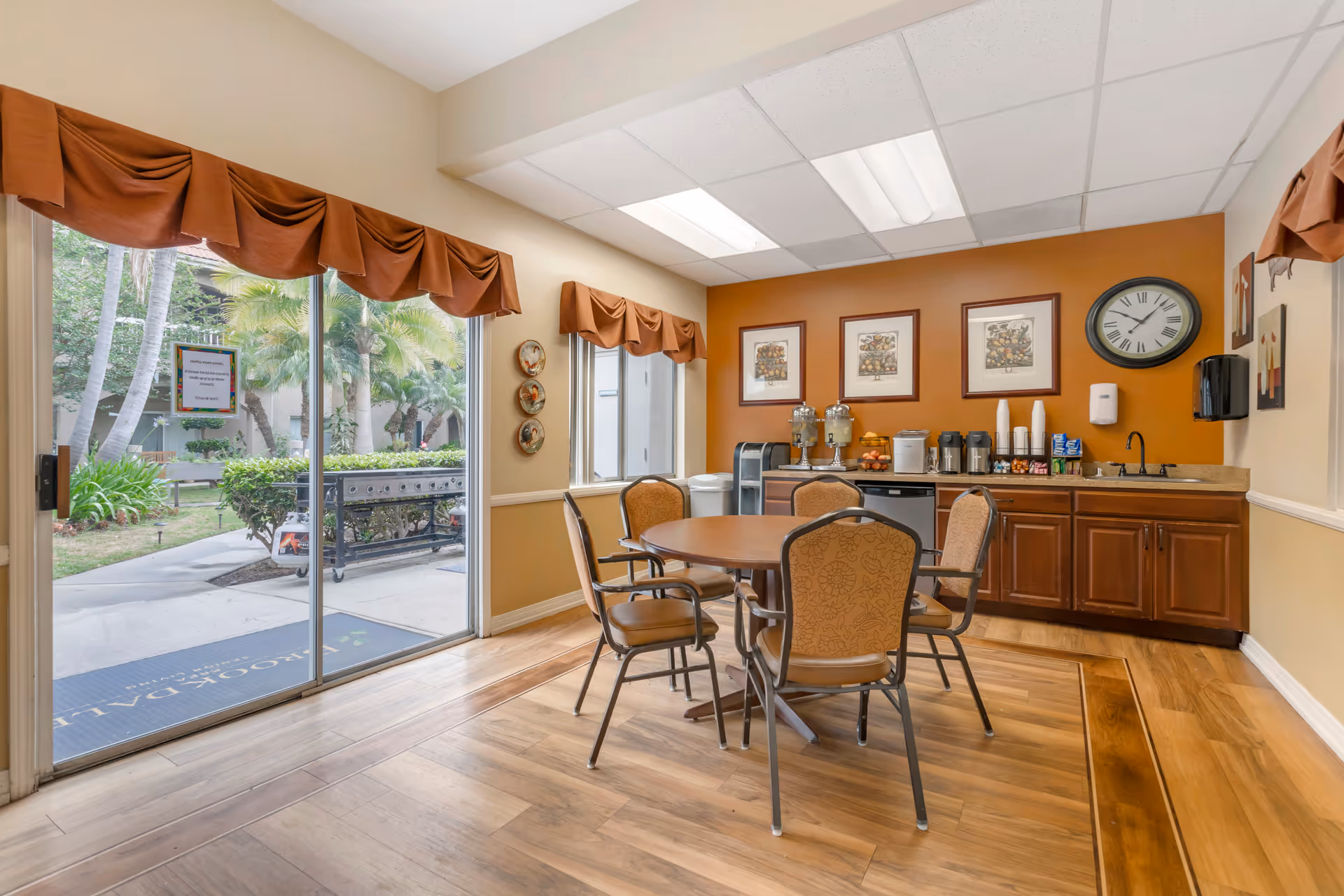 A small dining area with a round table and four chairs on a wooden floor. The room has a sliding glass door with a view of an outdoor garden area with palm trees. The back wall is painted orange and features three framed pictures, a clock, and a countertop with beverage dispensers, coffee machines, cups, and snacks. Brown valances adorn the windows and sliding door.