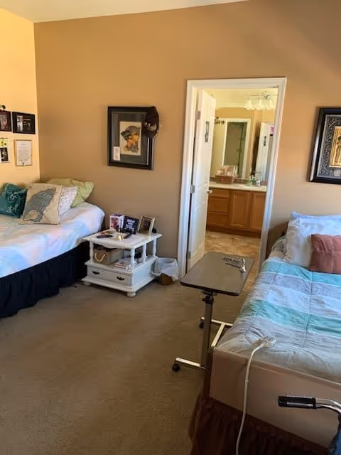 A cozy assisted living bedroom with two beds on opposite sides of the room. One bed has a white bedspread with decorative pillows, and the other has a striped bedspread with pillows. Between the beds is a small white nightstand with books and personal items. A door in the center of the back wall leads to a bathroom with a sink and mirror visible. The walls are painted beige, and framed artwork hangs on the walls.