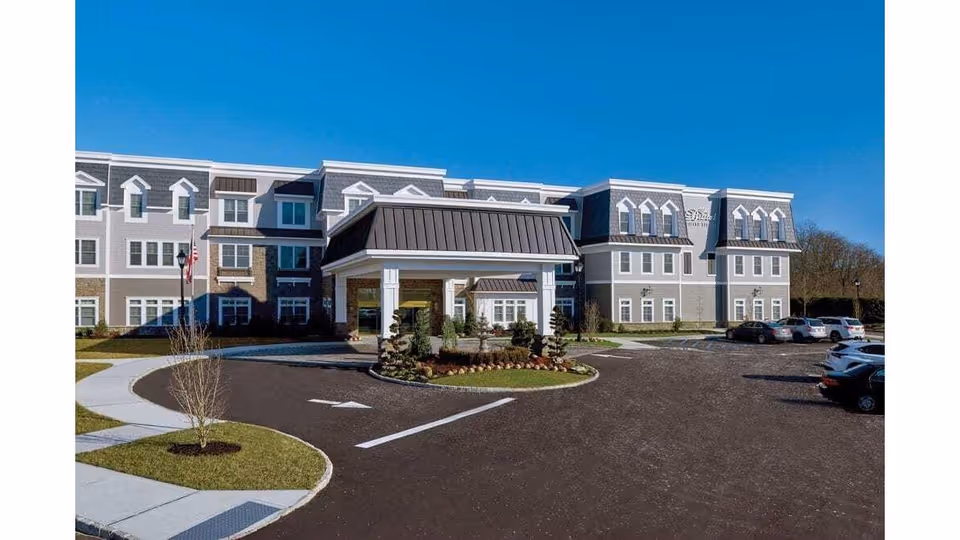 Exterior view of The Bristal Assisted Living at Jericho building with a driveway, landscaped entrance, and parked cars under a clear blue sky.