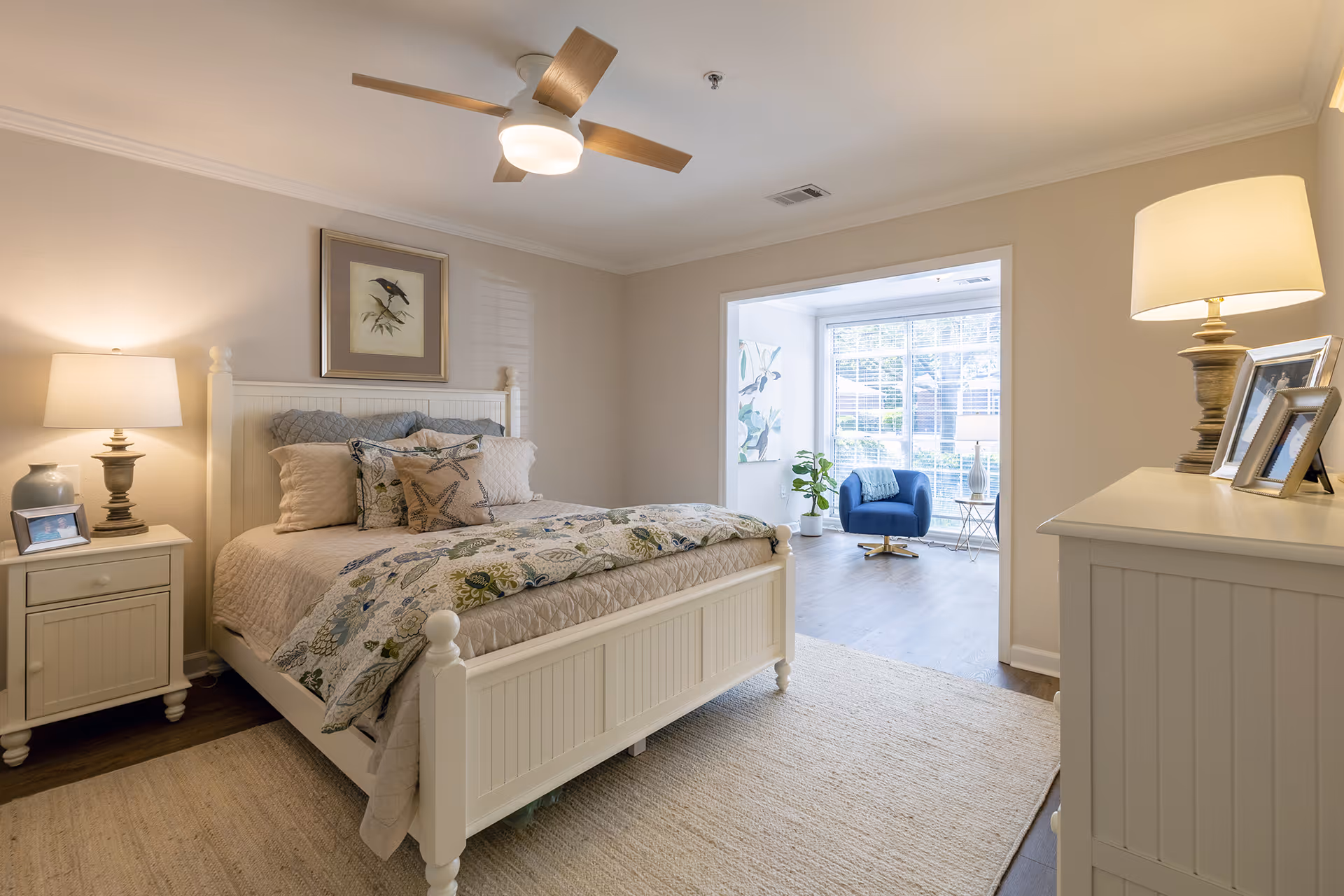Sunlit bedroom with a white wooden bed, bedside tables and a windowed sitting area with a blue chair.