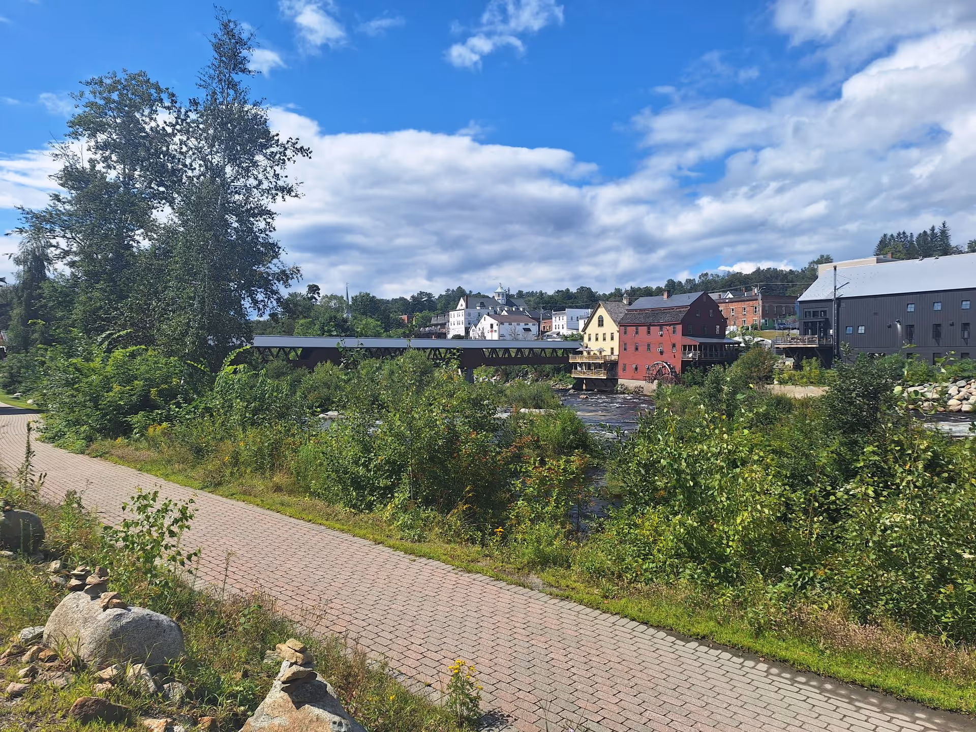 A scenic outdoor view featuring a paved walking path alongside a river with lush green vegetation. Across the river, there are several buildings including a red mill with a water wheel, a covered bridge, and other structures under a partly cloudy blue sky.