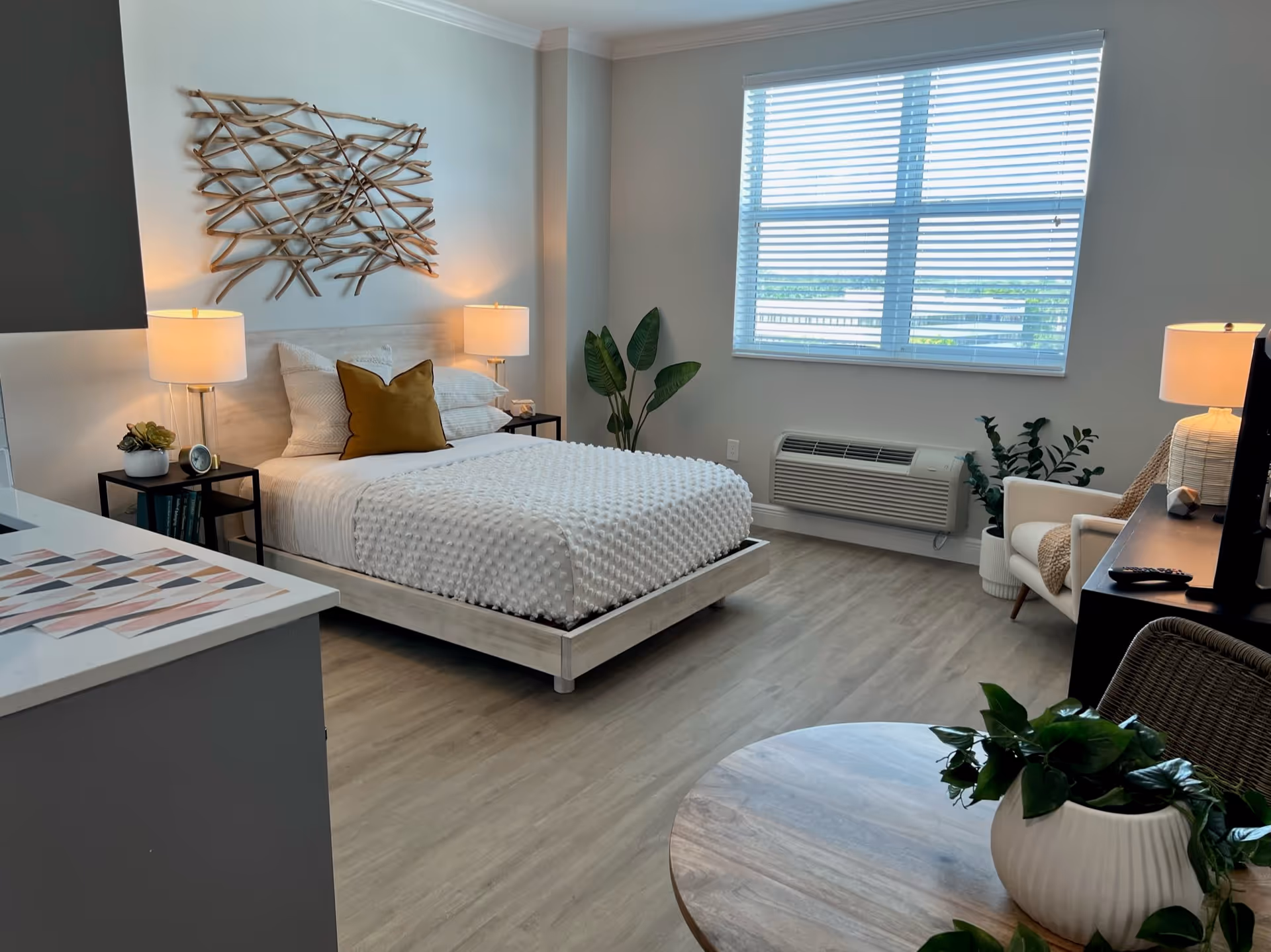 A bright and modern bedroom with a large window covered by white blinds. The room features a bed with white textured bedding and a mustard yellow pillow, two bedside tables with lamps, a decorative wooden wall art above the bed, a potted plant in the corner, a white armchair with a throw blanket, a TV on a dark stand, and a round wooden table with a green potted plant in the foreground.