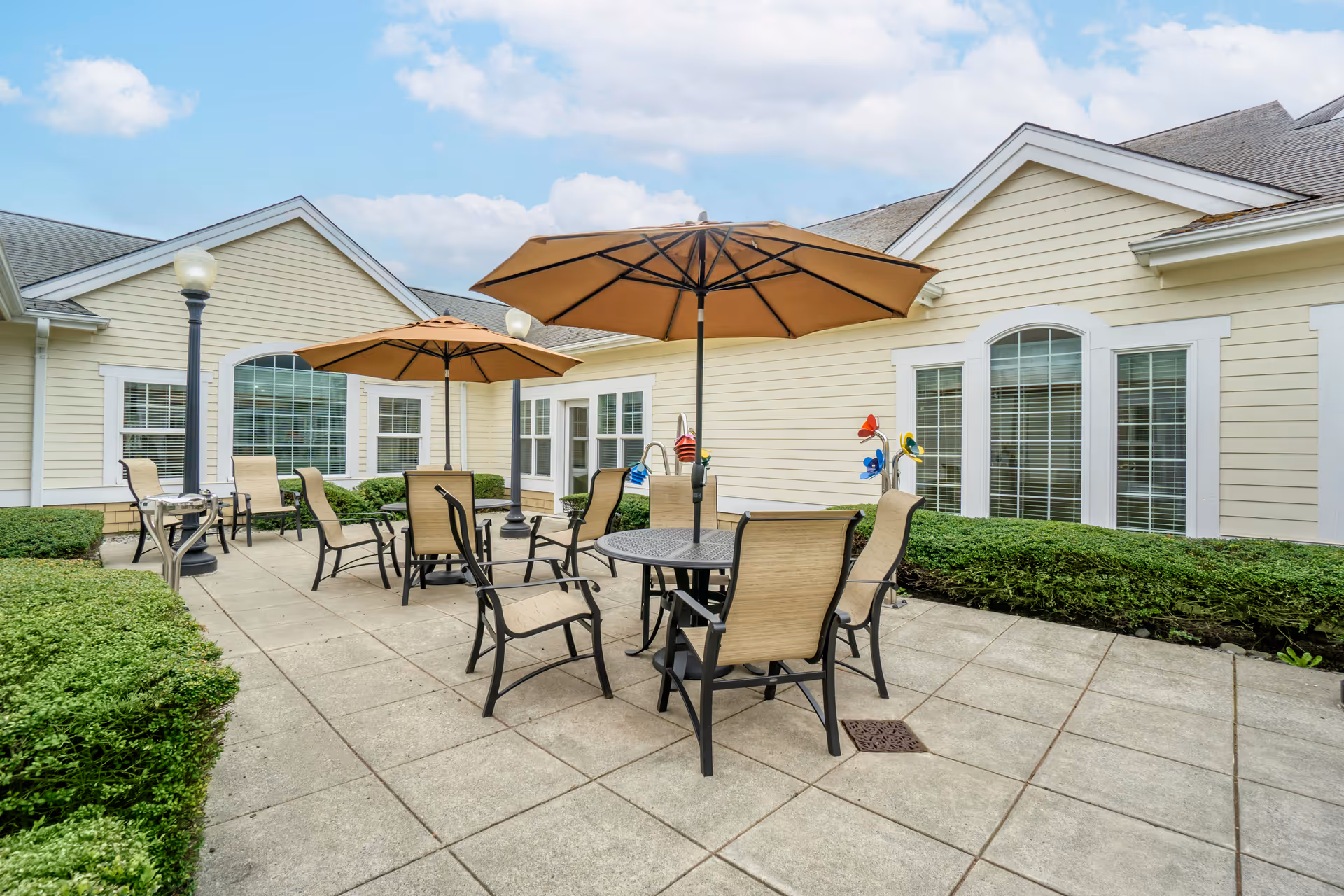 Outdoor patio area with several round tables and beige chairs under large brown umbrellas, surrounded by neatly trimmed bushes and light yellow building walls with white trim and windows.