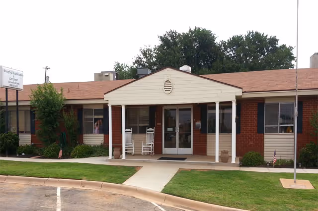 Front exterior view of Brownfield Rehabilitation and Care Center, a single-story brick building with a covered entrance supported by white columns. Two white rocking chairs are placed on the porch, and there is a small lawn with shrubs and an American flag near the entrance. Trees are visible in the background.