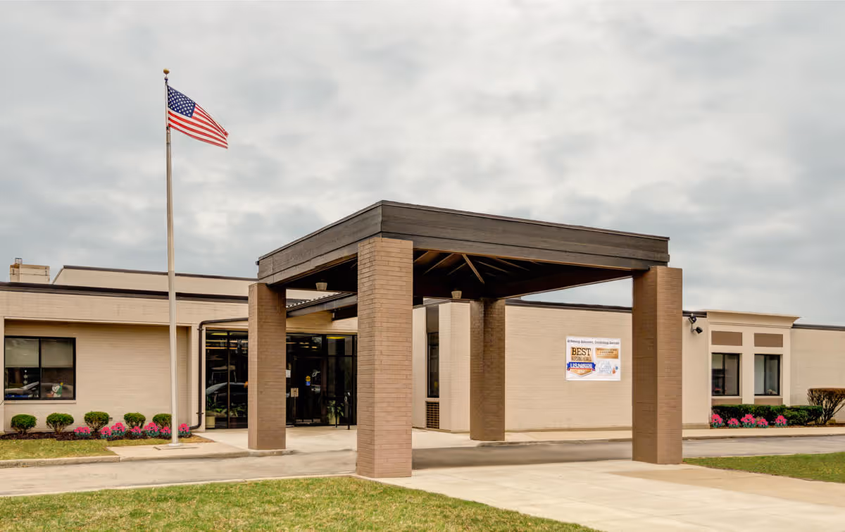 Exterior view of Oak Creek Terrace Nursing & Rehab Center showing the entrance with a covered drop-off area supported by brick pillars, an American flag on a flagpole, and a cloudy sky overhead. The building is light-colored with windows and some landscaping including bushes and flowers.