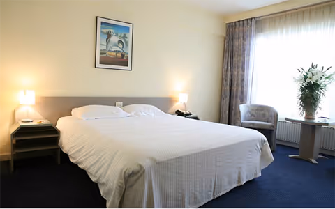 A neatly made bed with white linens in a well-lit bedroom featuring two bedside tables with lamps, a framed artwork on the wall above the bed, a window with sheer curtains, a patterned armchair, and a small round table with a vase of flowers.