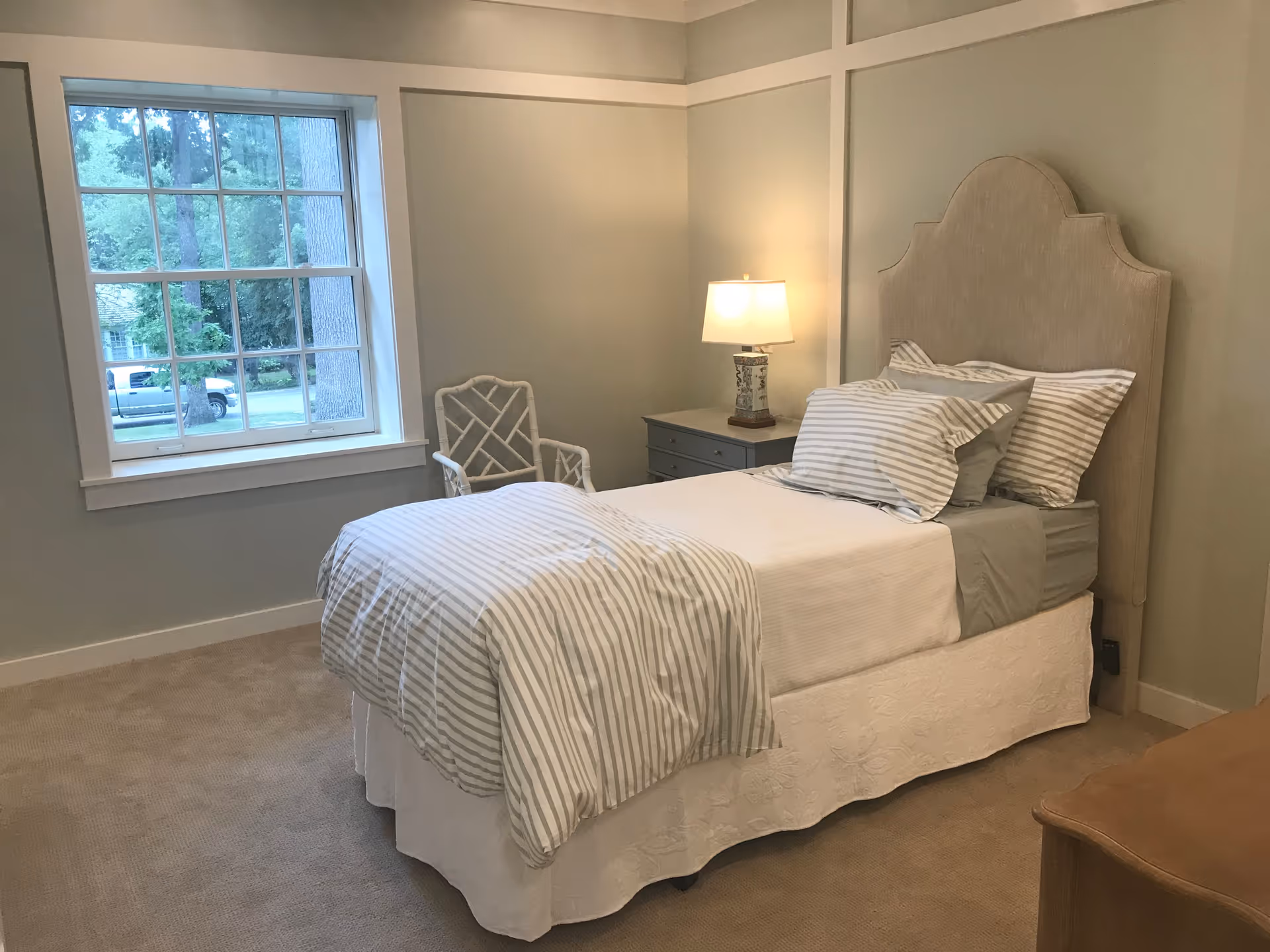 A neatly made single bed with striped bedding and multiple pillows in a softly lit bedroom. Next to the bed is a nightstand with a decorative lamp. A white chair is positioned near a large window showing a view of trees and a parked vehicle outside. The room has light-colored walls and carpeted flooring.