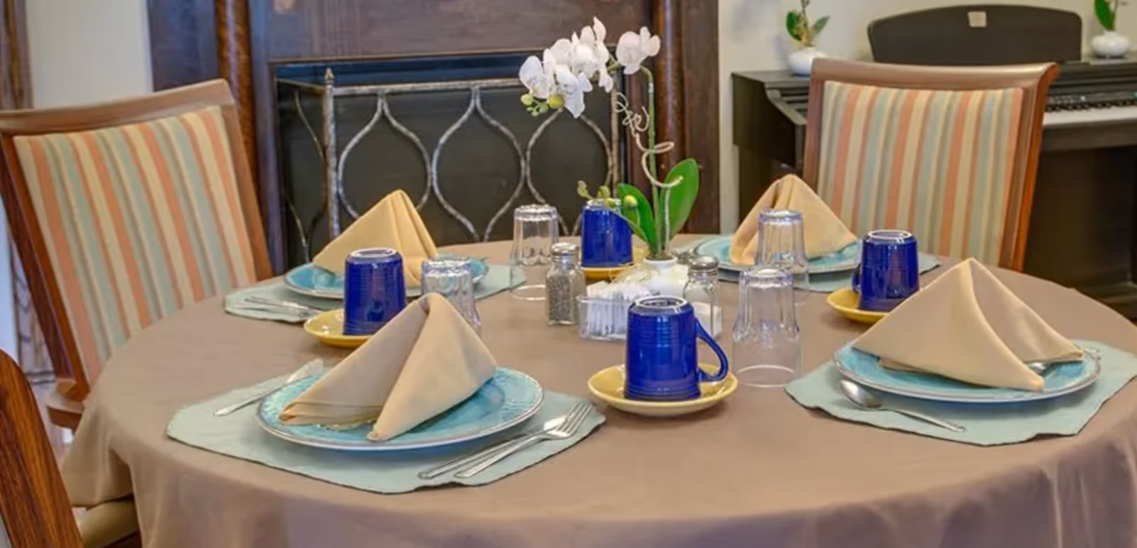 Round dining table set for four with plates, folded napkins, blue mugs and an orchid centerpiece in a cozy dining area.