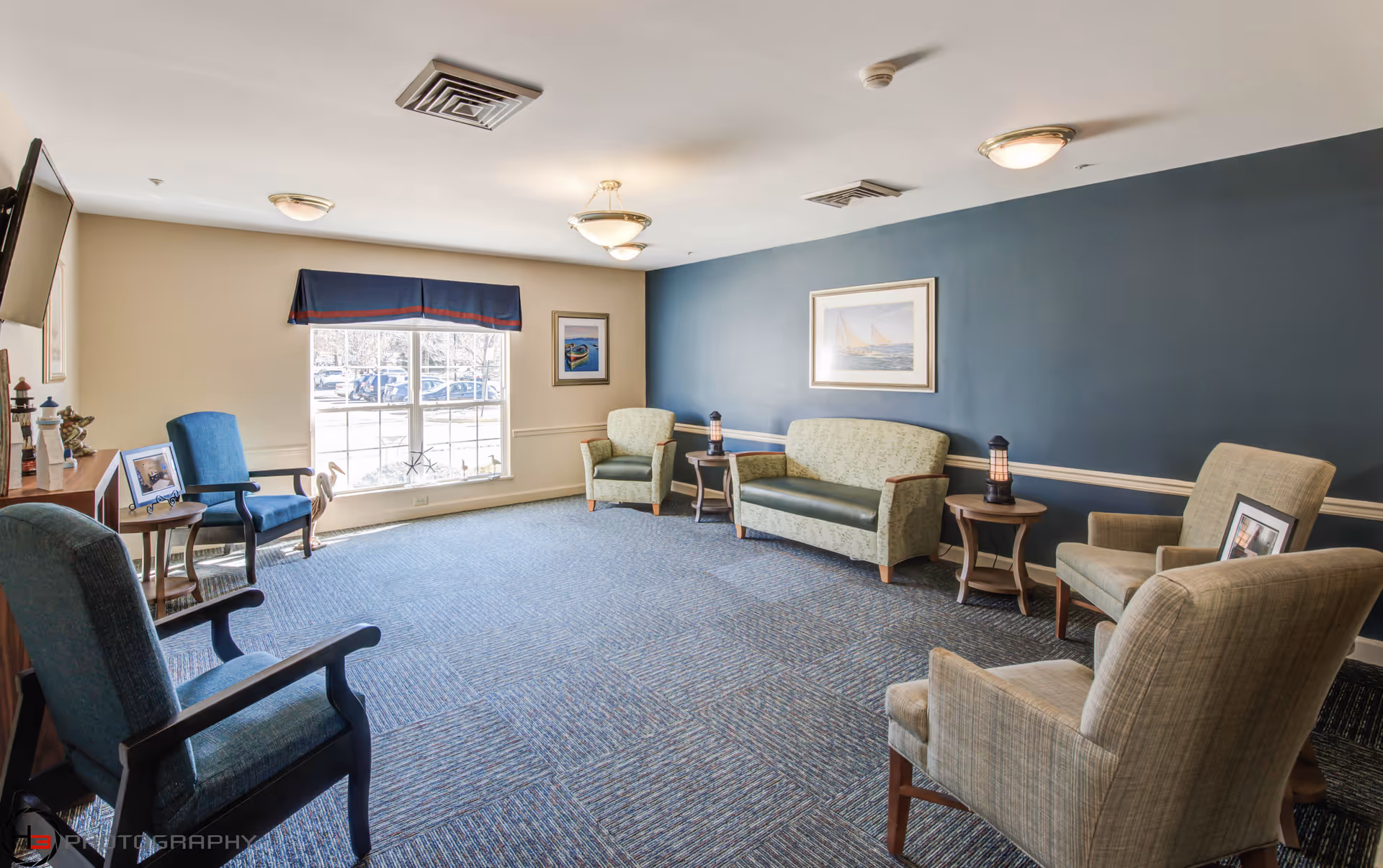 A bright communal sitting room with upholstered chairs, sofas, side tables and a large window.